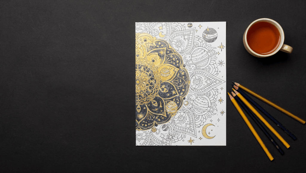 Coloring benefits: a half-colored cosmic mandala coloring page in gold and navy with colored pencils and a cup of tea on a black desk