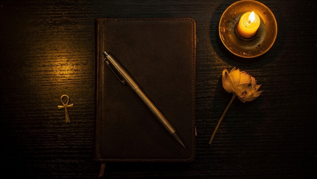 Past-life reflection practice: a closed leather journal with a pen, a candle, a dried lotus and an ankh on a dark desk